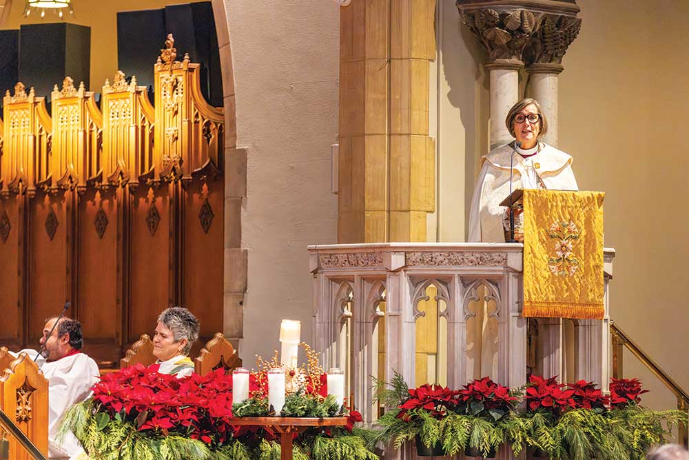 Archbishop Anne Germond standing in a stone pulpit adorned with a gold liturgical hanging, preaching during a church service decorated for Christmas with poinsettias and greenery, while other clergy sit below.