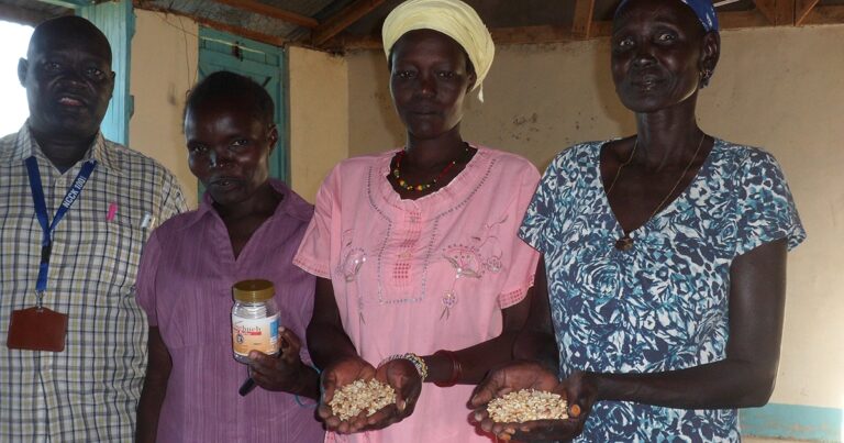 Three women holding containers of food and grains in a community setting, showcasing local agricultural products and food security efforts.