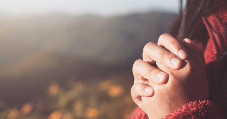 A person’s hands clasped together in prayer, set against a softly blurred outdoor background. The individual wears a red sweater, and the warm light suggests early morning or late afternoon.