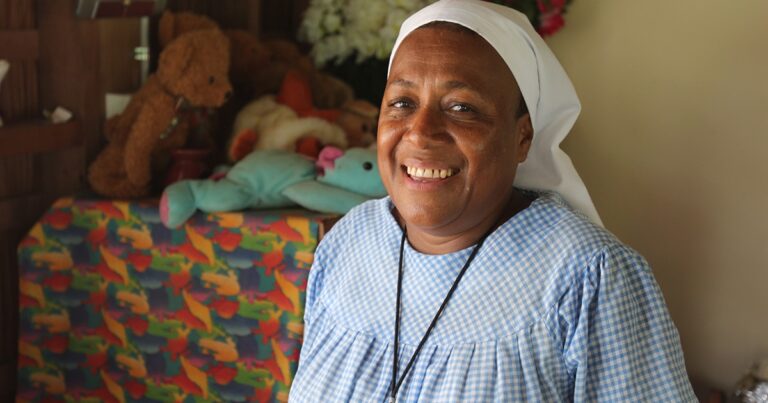 Smiling woman wearing a blue dress and white headscarf, standing in front of a colorful background with stuffed animals.