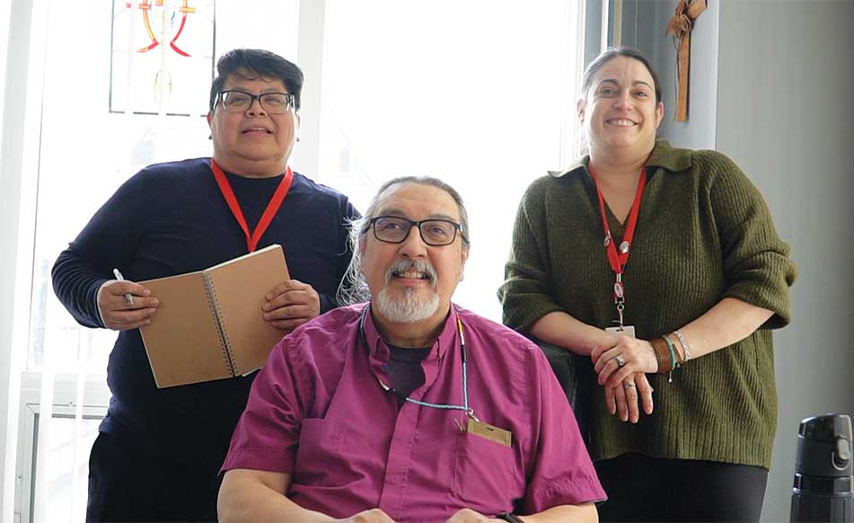 Three people indoors smiling toward the camera: an older man seated at the centre wearing glasses and a magenta clergy shirt, and two colleagues standing on either side of him with notebooks and ID lanyards.