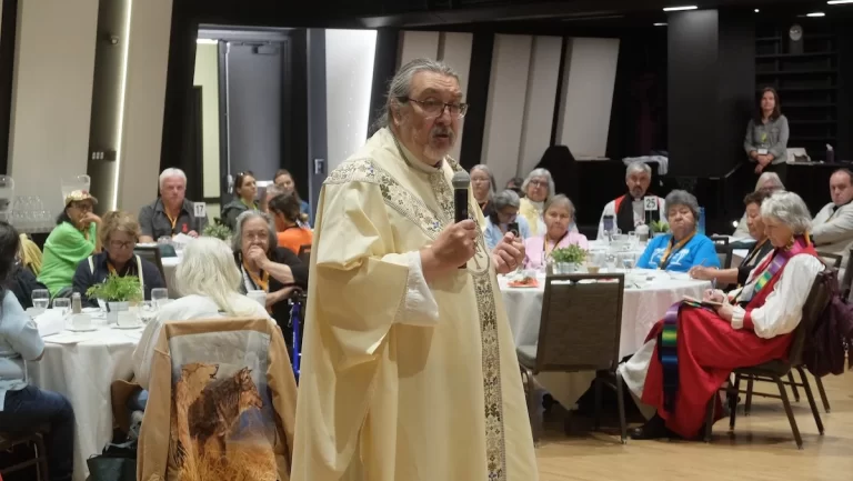 Archbishop Chris Harper speaks into a microphone at Sacred Circle, addressing Indigenous clergy and community members seated at round tables. He wears a cream-coloured chasuble adorned with detailed gold and blue embroidery along the edges.