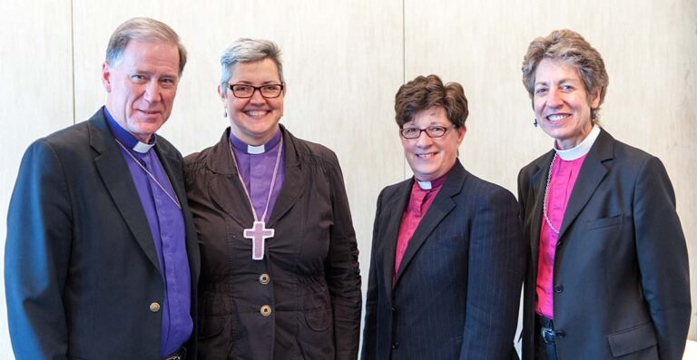 A group of four clergy members, including two women and two men, standing together and smiling. The woman in the center wears a clerical collar and a cross necklace, while the others are dressed in formal attire. The background is a light-colored wall, indicating a professional or ceremonial setting.