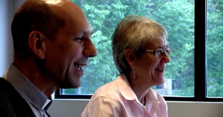 Two adults engaged in conversation, smiling and appearing to enjoy a discussion, with a green outdoor background visible through a window.