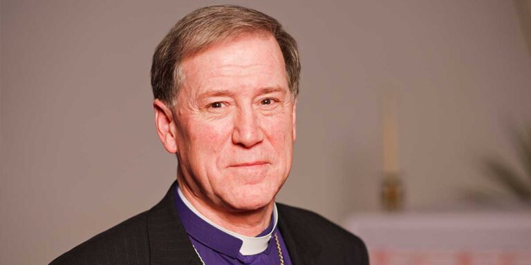 A close-up portrait of a man with short, light brown hair and a slight smile, wearing a black suit and clerical collar, set against a neutral background.