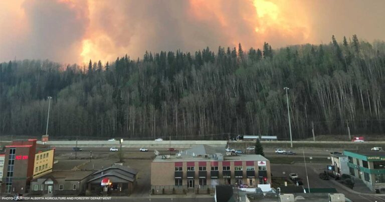 Wildfire smoke billowing over a forested area, with a hotel visible in the foreground, highlighting the impact of environmental events on local communities.