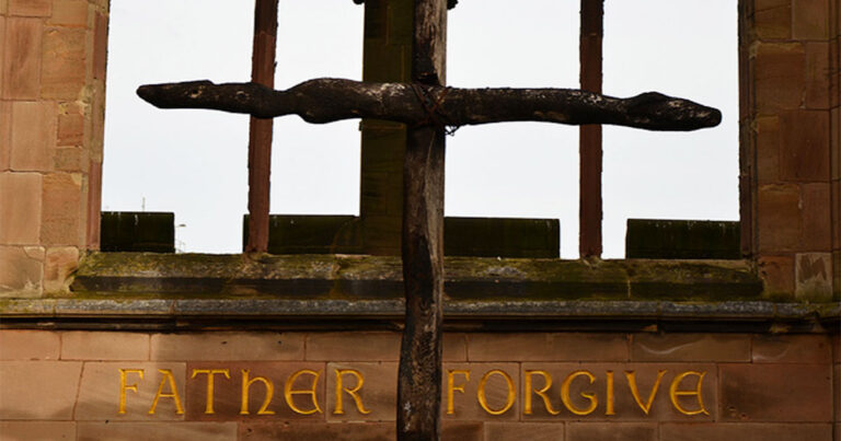 Close-up of a wooden cross structure with the words "Father Forgive" inscribed below, set against a cloudy sky background.