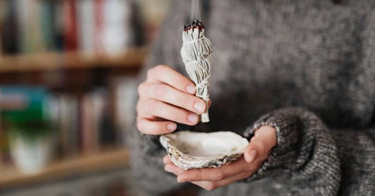 A person holding a bundle of sage above a small shell dish, preparing to perform a smudging ritual. The setting has a cozy, natural aesthetic, with blurred background elements.