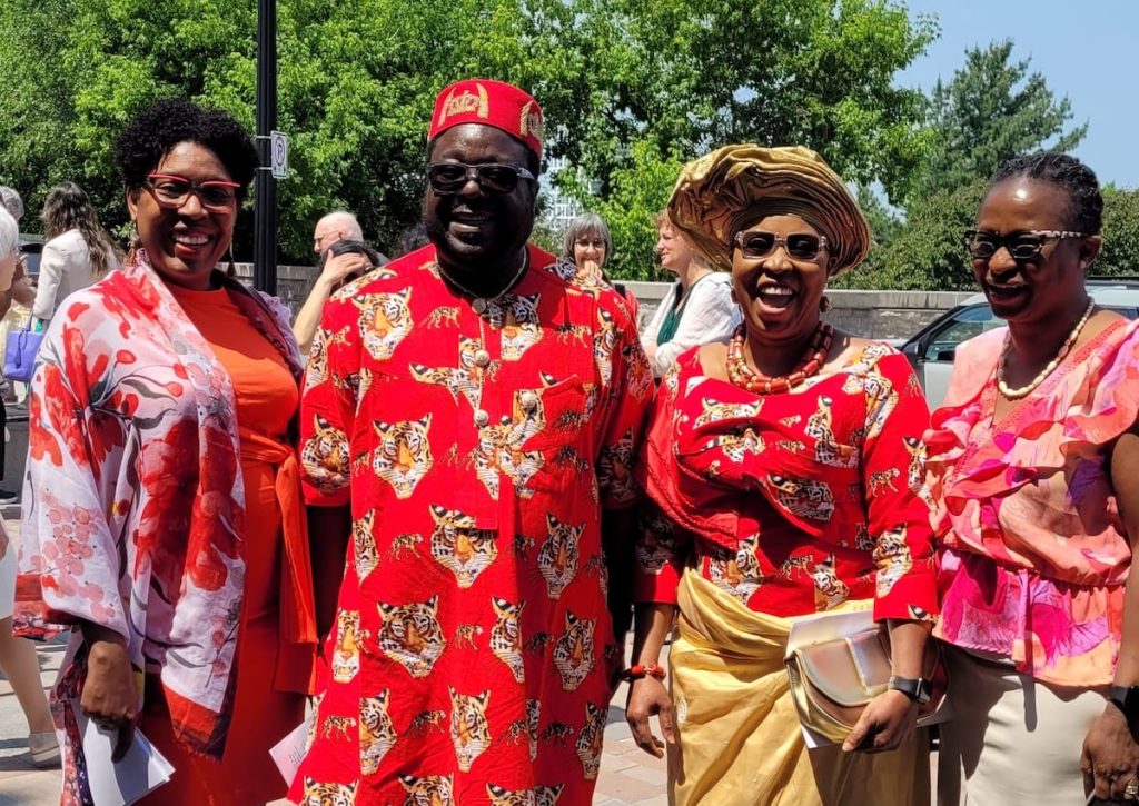 Four people wearing brightly coloured red and pink traditional clothing smile together outdoors during a community celebration or church gathering