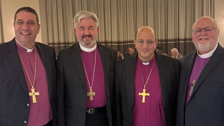 Four Anglican and Evangelical Lutheran bishops—Archbishop Hosam Naoum, Archbishop Shane Parker, Bishop Imad Haddad, and Bishop Larry Kochendorfer—stand side by side indoors, wearing clerical shirts with pectoral crosses, at a gathering in Jerusalem for Bishop Haddad’s consecration.