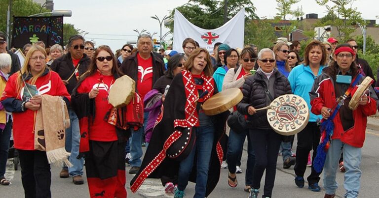 A group of diverse individuals marching together, carrying drums and wearing traditional clothing, in a public demonstration celebrating Indigenous culture and heritage.