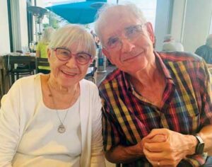 Older couple smiling at the camera while seated in a bright café or restaurant; the woman wears glasses and a white top, and the man wears glasses and a multicoloured plaid shirt.