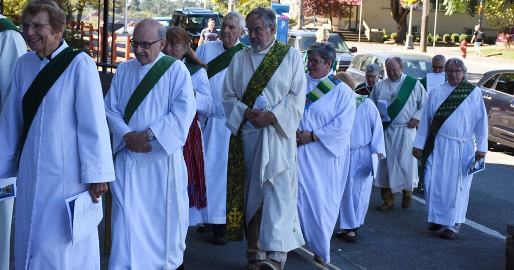 A group of clergy members in white robes and green stoles walking in a procession along a street, with a crowd and vehicles in the background.