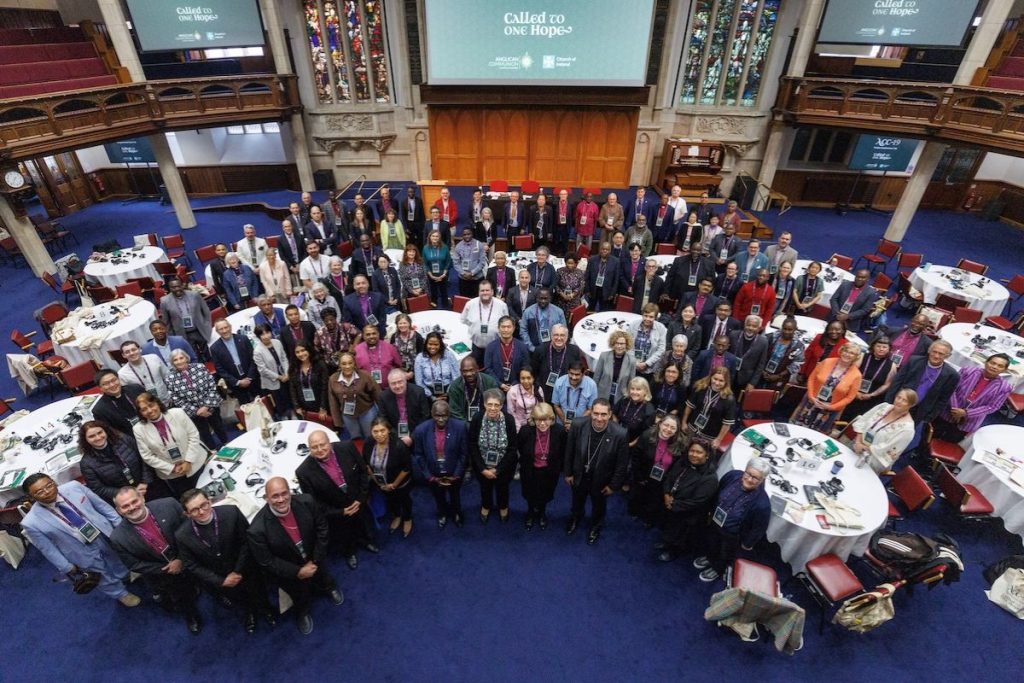 A diverse group of attendees, including bishops in purple attire, stands in a semi-circle in a blue-carpeted room with round tables, facing the camera. A large screen in the background displays "CALLED to ONE Hope." The 19th Anglican Consultative Council (ACC-19) gathers for a group photo at the end of their June 28 morning session at the Assembly Buildings Conference Centre in Belfast, Northern Ireland. Photo: Neil Turner for ACO