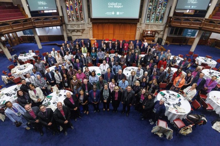 A diverse group of attendees, including bishops in purple attire, stands in a semi-circle in a blue-carpeted room with round tables, facing the camera. A large screen in the background displays "CALLED to ONE Hope." The 19th Anglican Consultative Council (ACC-19) gathers for a group photo at the end of their June 28 morning session at the Assembly Buildings Conference Centre in Belfast, Northern Ireland. Photo: Neil Turner for ACO