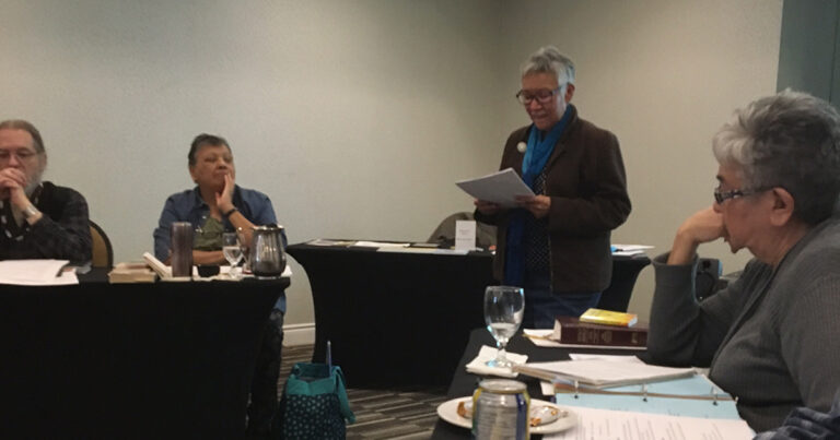 A speaker stands in front of an audience during a meeting, holding notes while discussing a topic. Two attendees are seated at a table, with various items like glasses and a can visible.