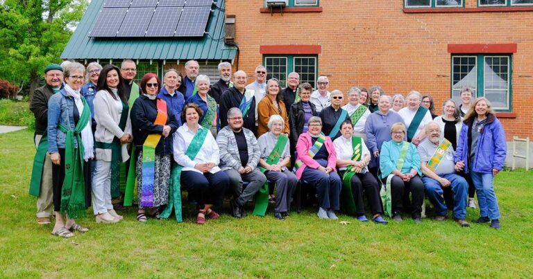 Group photo of a diverse gathering of individuals wearing colorful sashes, standing and sitting in front of a brick building with solar panels on the roof, surrounded by greenery.