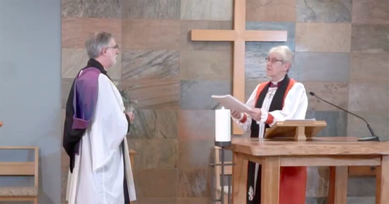 A clergy member in white robes stands before another clergy member in a church setting, with a cross in the background. The first clergy member is holding a document, while the second appears to be listening attentively.