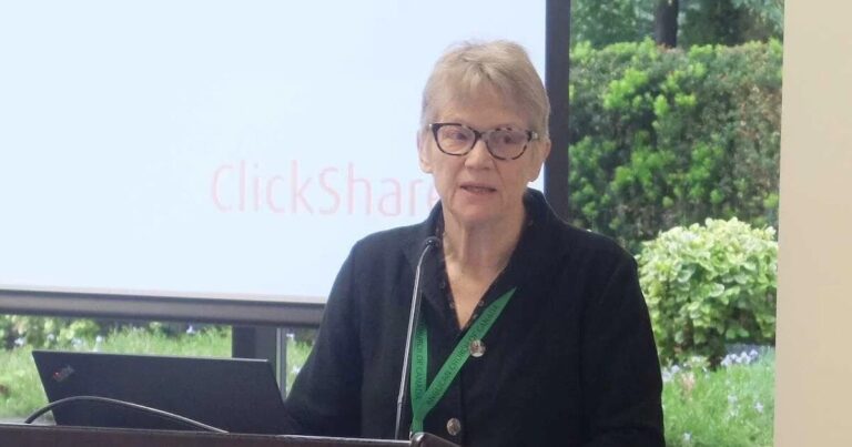 An older woman with short light hair and glasses stands at a podium, speaking into a microphone. She wears a dark jacket and a green conference lanyard. Behind her, a presentation screen displays the word “ClickShare,” and greenery is visible through the windows.