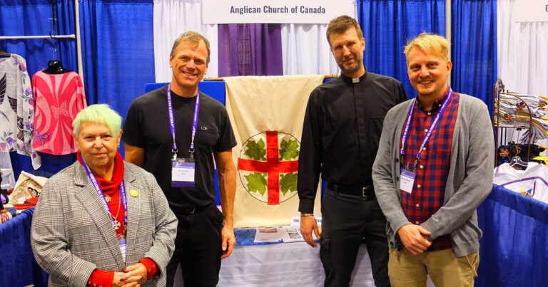 Group of four individuals standing in front of a display for the Anglican Church of Canada, featuring a banner with a red cross and greenery. The setting appears to be a conference or exhibition.