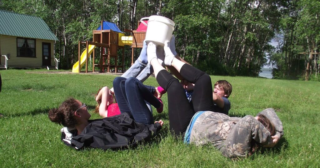 Group of children playing a game on a grassy field, with one child holding a bucket above their feet while others lay on their backs, laughing and engaging in the activity. In the background, a playground structure is visible.