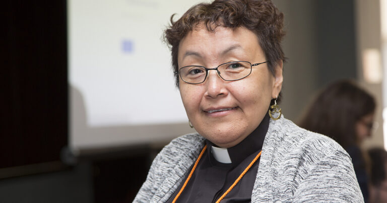 Portrait of a smiling woman with short, curly hair, wearing glasses and a clerical collar, seated in front of a blurred background. She appears confident and approachable, emphasizing a warm expression.