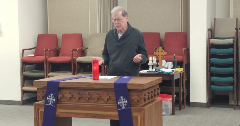 Elderly man standing at a wooden altar with a red candle and purple cloth, speaking in a church setting with chairs in the background.
