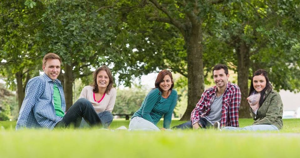 A group of three young adults sitting on grass in a park, smiling and enjoying a sunny day, surrounded by trees.