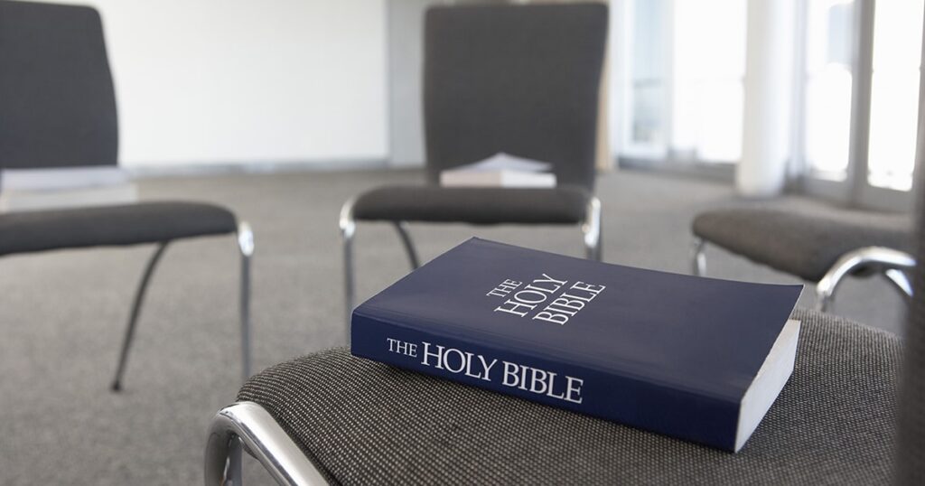 A close-up of a blue-covered Holy Bible resting on a gray chair, with a blurred background of additional chairs and papers.