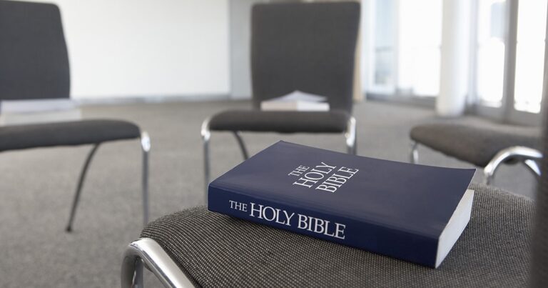 A close-up of a blue-covered Holy Bible resting on a gray chair, with a blurred background of additional chairs and papers.
