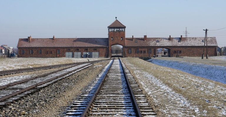 Auschwitz concentration camp entrance with train tracks leading towards the gate, surrounded by barren landscape and snow.