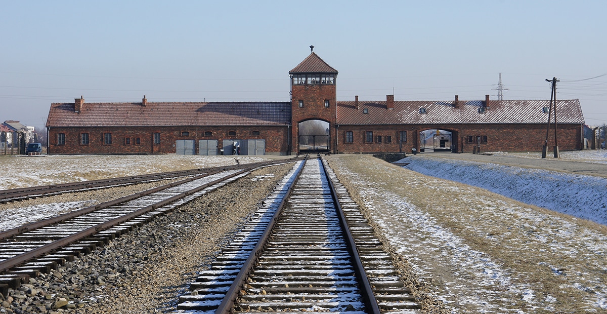 Auschwitz concentration camp entrance with train tracks leading towards the gate, surrounded by barren landscape and snow.