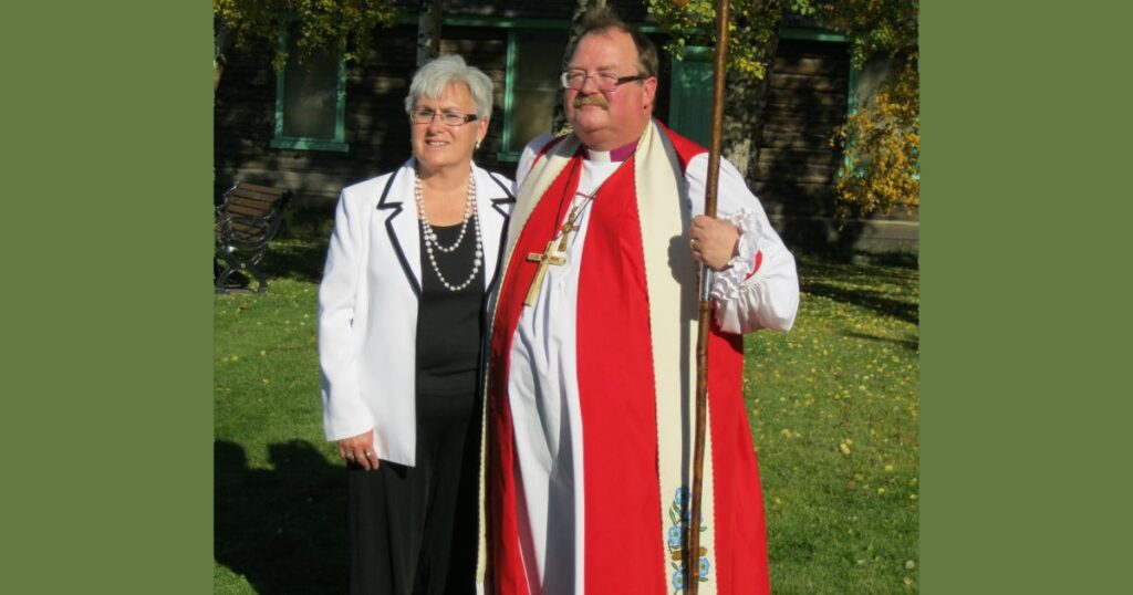 A man wearing a red ceremonial outfit and holding a staff stands next to a woman in a black dress and white blazer, smiling in a green outdoor setting.