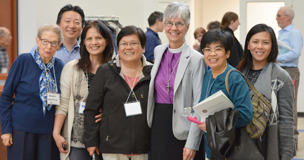 Group of five diverse women smiling together at an event, showcasing camaraderie and engagement. The setting appears to be a conference or gathering, with attendees in the background.