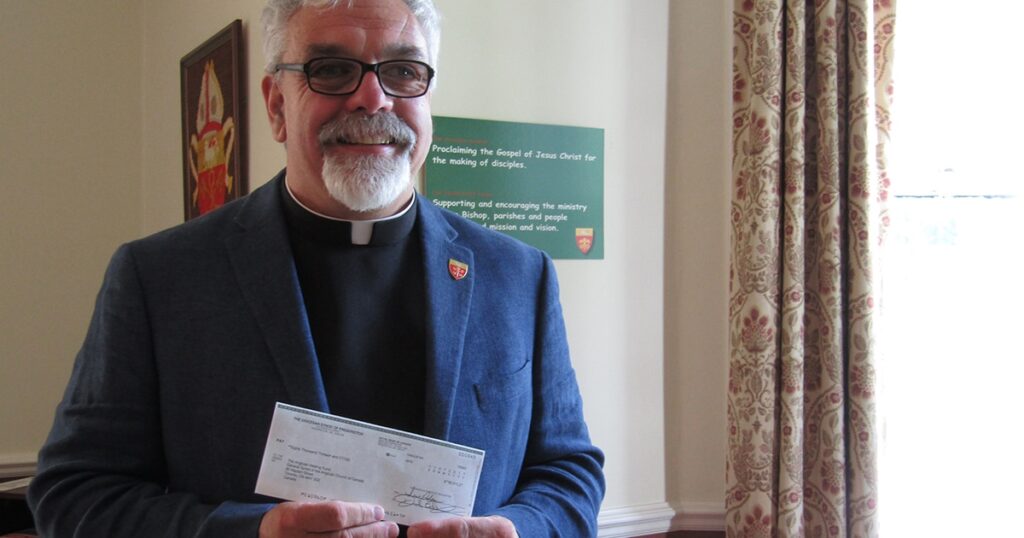 A man in a dark blazer and clerical collar holds a check in front of a green sign in a well-lit room.