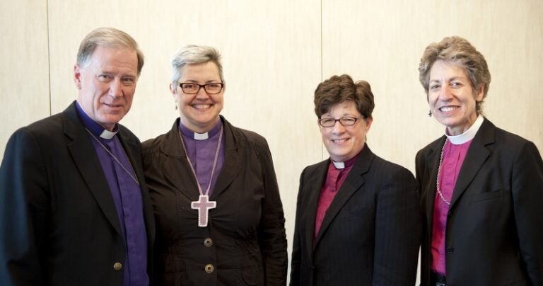 Three clergy members pose together, smiling, in a professional setting. They are dressed in formal attire, with one wearing a clerical collar and a cross necklace. The background is neutral and minimalistic, emphasizing the individuals in the image.
