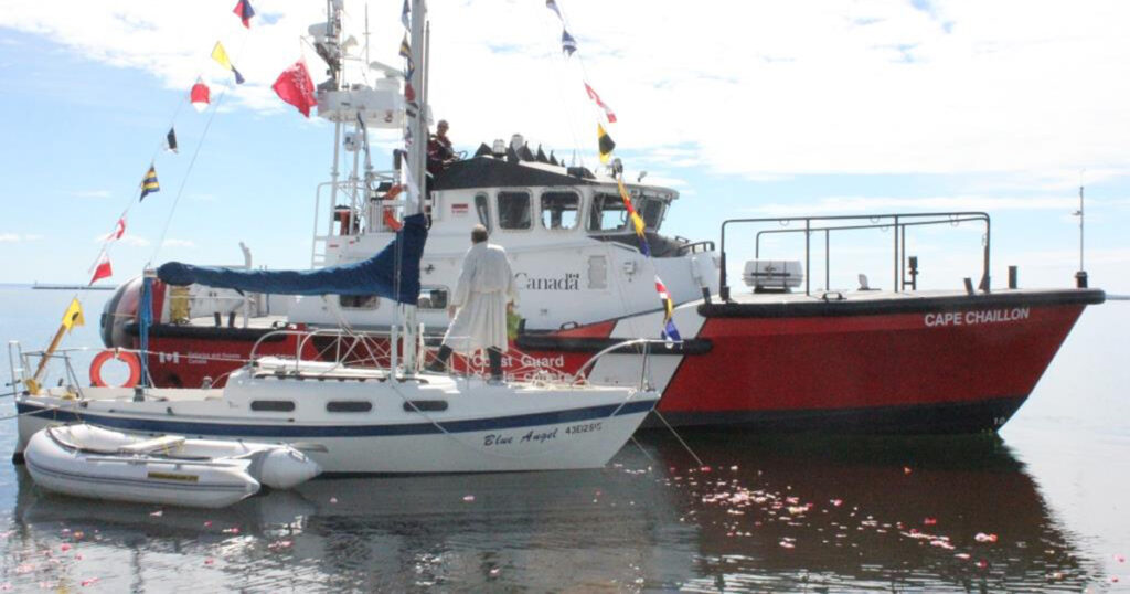 Sailboat anchored near a large tugboat, both decorated with colorful flags, on a calm water surface under a clear sky.
