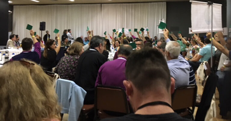 A crowded conference room with attendees raising green cards in a voting process, indicating participation and decision-making during a meeting.