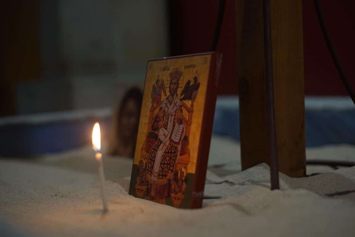 A candle lit beside a religious icon featuring a figure surrounded by symbolic elements, placed on a textured surface. The background is softly blurred, enhancing the focus on the icon.