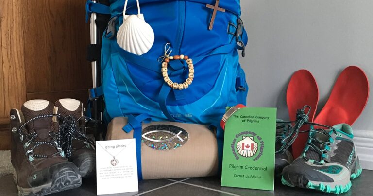 A blue backpack adorned with a seashell, a decorative cross, and a beaded keychain, accompanied by a rolled-up mat and two pamphlets, one green and one white, on a tiled surface.