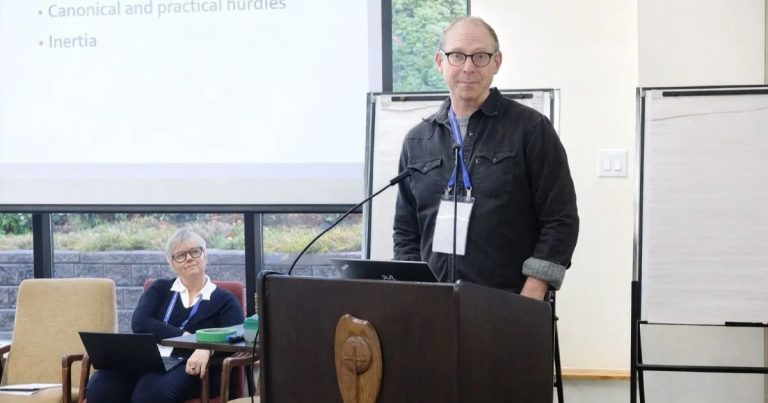 Transformation Task Force co-chair Canon (lay) Andrew Stephens-Rennie stands at a podium, wearing a black jacket with glasses.