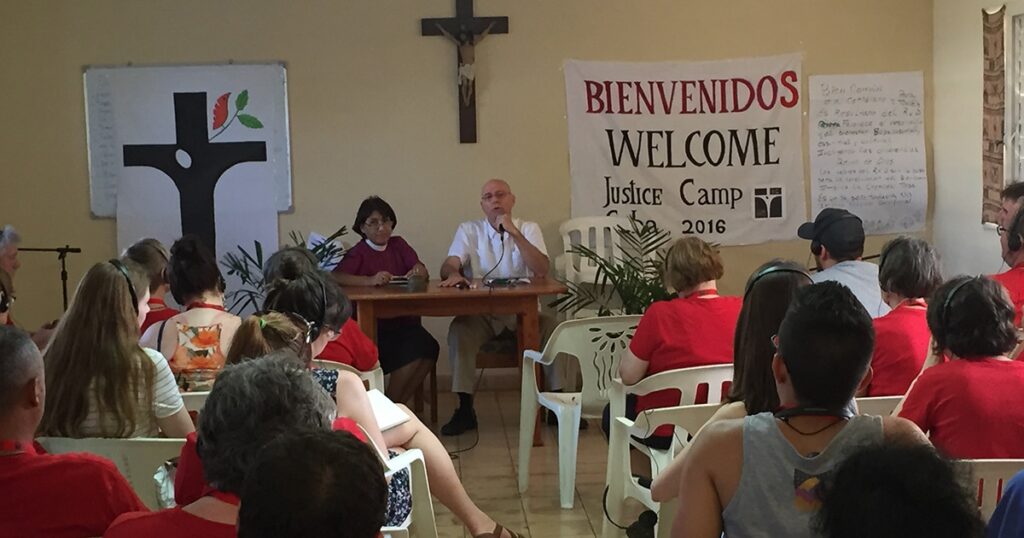 Panel discussion at a welcome event for a justice camp, featuring two speakers at a table in front of a banner reading "Bienvenido" and "Welcome," with an audience seated in white chairs. A cross is visible in the background.