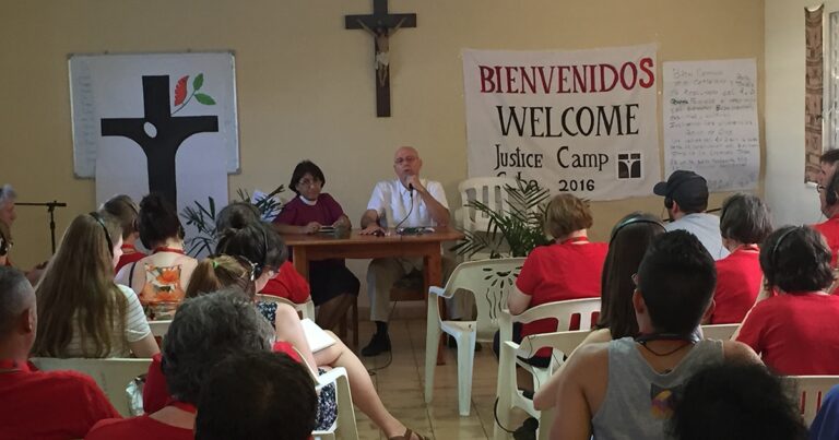 Panel discussion at a welcome event for a justice camp, featuring two speakers at a table in front of a banner reading "Bienvenido" and "Welcome," with an audience seated in white chairs. A cross is visible in the background.