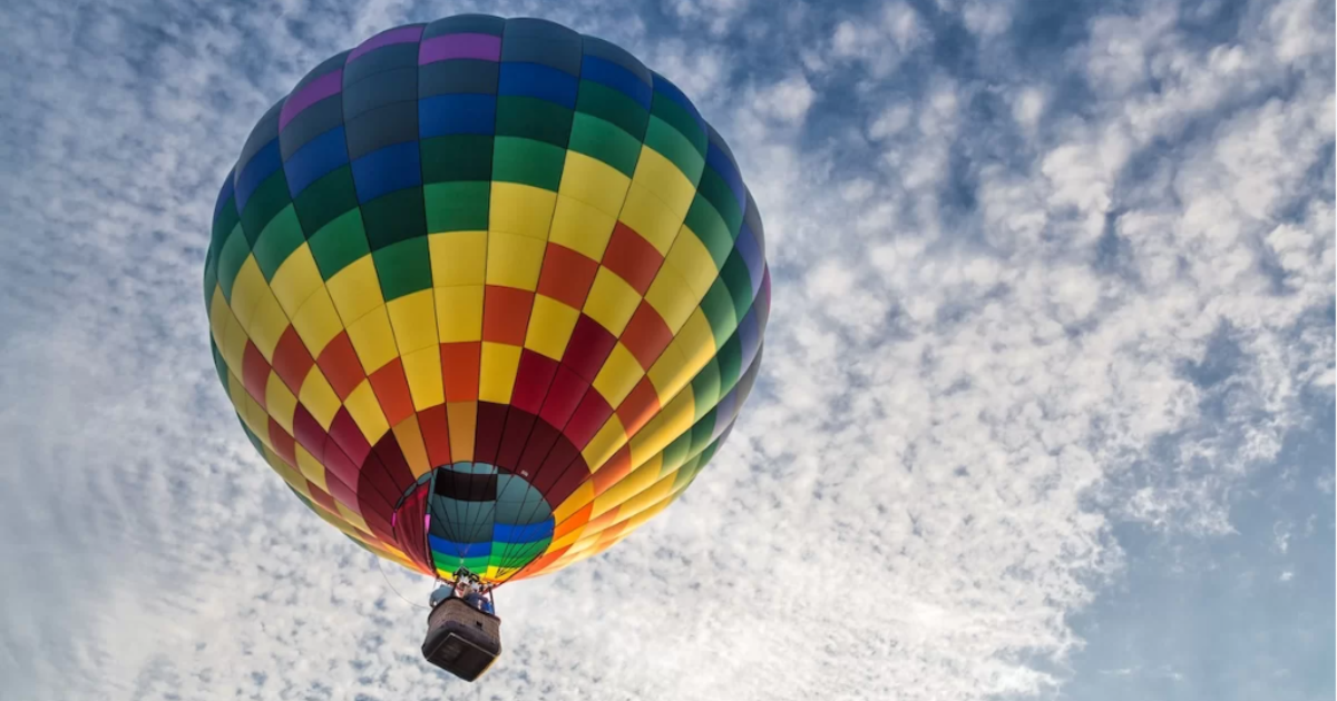 Image shows a large hot air balloon that is rainbow-coloured, flying high against the backdrop of a cloudy, blue sky.