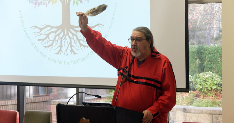 A man in a red traditional outfit raises a feather while speaking at a podium, with a tree roots logo visible in the background.