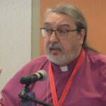 A speaker with long hair and glasses gestures while addressing an audience at a conference, wearing a purple clerical shirt and a red lanyard.