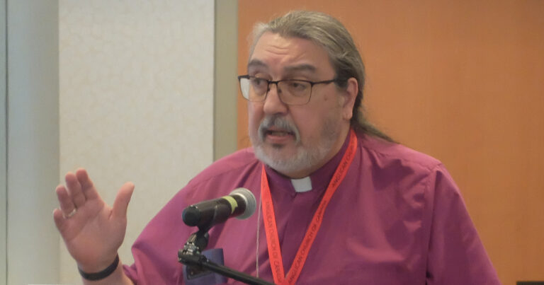 A speaker with long hair and glasses gestures while addressing an audience at a conference, wearing a purple clerical shirt and a red lanyard.