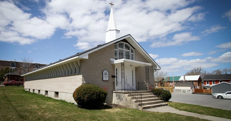 A small church building with a white steeple, surrounded by green grass and shrubs, under a partly cloudy sky.