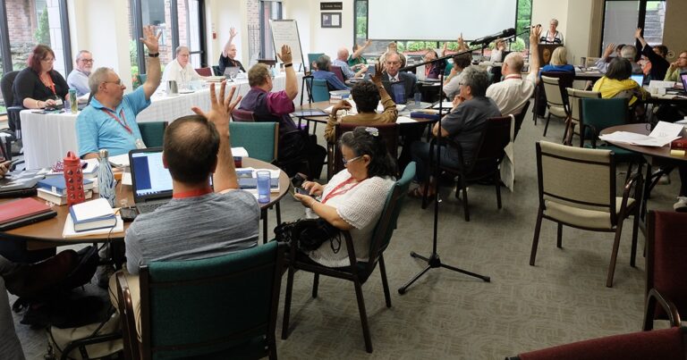 A group of people sitting in a conference room, raising their hands during a discussion, with tables set up for an interactive meeting.