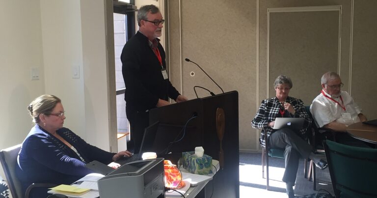 A speaker presenting at a podium during a conference, with an audience member seated nearby, engaged in note-taking.